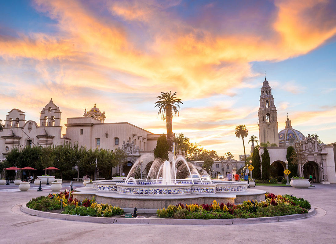 Contact - Flowers Around a Fountain with Palm Trees and Historical Buildings in the Background Against a Colorful Sunset Sky